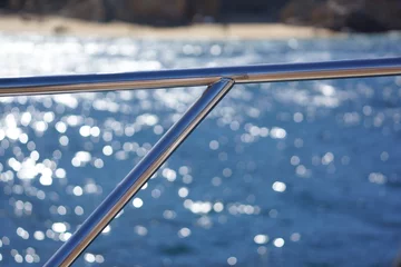 Schilderijen op glas Macro of shiny metal rail on boat, ocean and rocky beach in background with bokeh © Eni