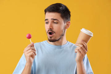 Handsome shocked young man with lollipop and cup of coffee on yellow background. Dental care concept