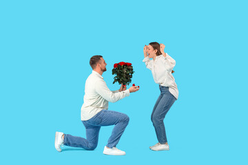 Young man with bouquet of roses proposing to his girlfriend on blue background. Valentine's Day celebration