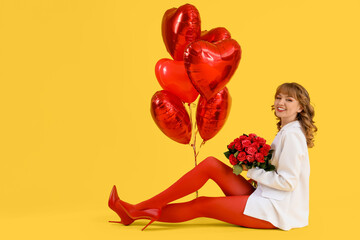 Beautiful young woman with bouquet of red roses and heart shaped air balloons on yellow background. Valentine's Day celebration