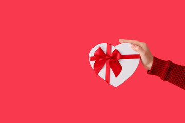 Female hand holding heart-shaped gift box on red background. Valentine's Day celebration