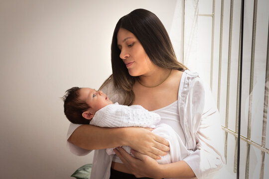 Mam&aacute; sonriendo cargando a su beb&eacute;, ambos vestidos de blanco, en el interior de su casa iluminados con luz natural