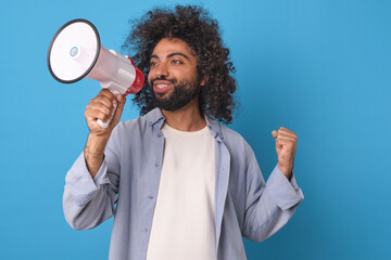 Young positive casual Arabian man student makes victory gesture and holds megaphone informing others about opportunity to go to college for free thanks to educational grant stands on blue background.