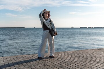 Stylish seashore woman. Fashionable woman in a white hat, white trousers and a light sweater with a black pattern on the background of the sea.
