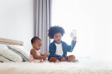 Happy African siblings in family, cute toddle baby infant with headphones listening music, brother boy with curly hair play with his sister girl on white bed at bedroom, kid child spend time together.