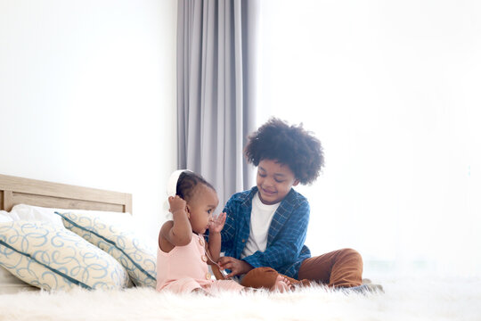 Happy African siblings in family, cute toddle baby infant with headphones listening music, brother boy with curly hair play with his sister girl on white bed at bedroom, kid child spend time together.