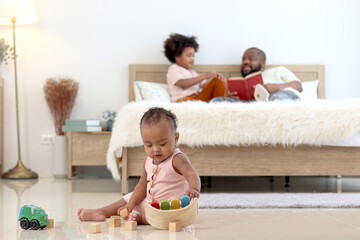 Obraz na płótnie Canvas African little cute toddle baby infant kid plays toy while sits on floor in home bedroom with blurred background of father and brother read book in bed. Happy family spending time together in bedroom.