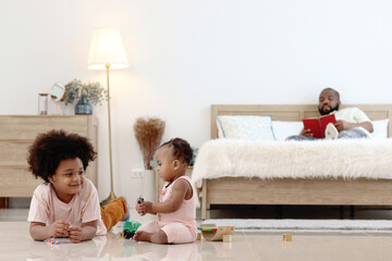 Obraz na płótnie Canvas Happy African family spending time together, brother boy with black curly hair lying on floor, playing toy with little cute toddle baby infant kid in bedroom. Sibling relationships in childhood.