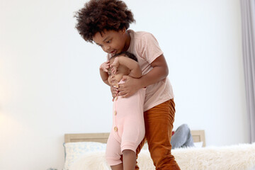 Happy African family spending time together, brother boy with black curly hair try to hold and lift up little cute toddle baby infant kid on floor with blurred background of dad reading book on bed.