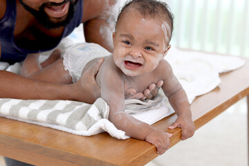 Happy African family, toddle baby infant lying on white towel after taking a bath and apply talcum...