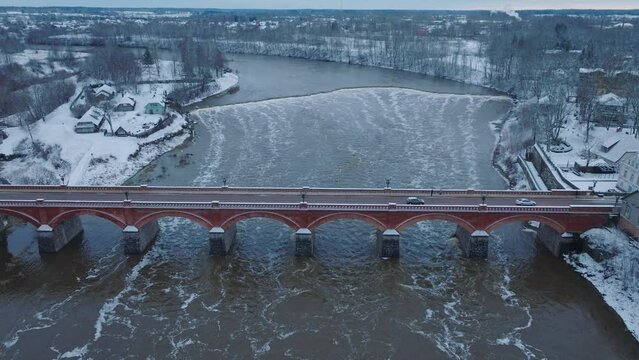 Aerial establishing view of Venta river rapids (Ventas rumba) during winter flood, old red brick bridge, Kuldiga, Latvia, overcast winter day, wide birdseye drone shot moving forward