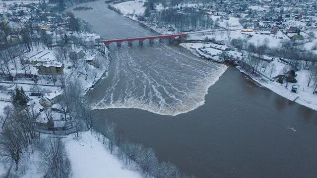 Aerial establishing view of Venta river rapids (Ventas rumba) during winter flood, old red brick bridge, Kuldiga, Latvia, overcast winter day, wide drone shot moving forward tilt down