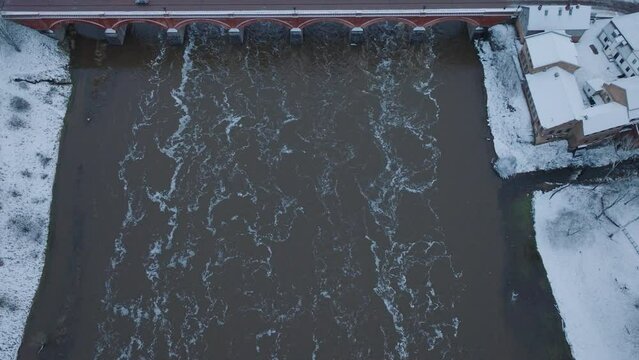 Aerial establishing view of Venta river rapids (Ventas rumba) during winter flood, old red brick bridge, Kuldiga, Latvia, overcast winter day, wide birdseye drone shot moving forward
