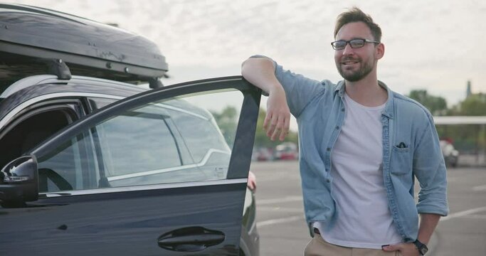 Young Caucasian man standing near the car with roof racks and cargo box on a parking lot and looking at camera