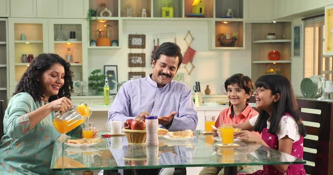 Happy Indian family mother pouring juice and father spreading jam on toast for kids
