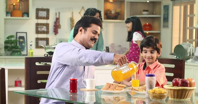 Video of father pouring orange juice to son and spreading jam on bread during breakfast while sitting on table in kitchen 
