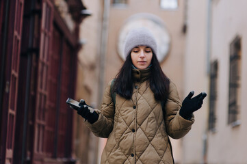 A young girl in a khaki down jacket and an angora hat with a smartphone spreads her hands against...