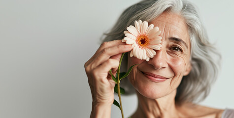 Photo portrait of senior 60 years old woman with fine lines and grey hair smiling looking at camera holding a flower next to her face, horizontal photo with copy space for banner