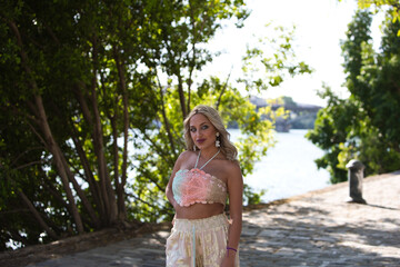 Young beautiful blonde woman dressed in embroidered trousers and crochet top by the river bank in seville. In the background green branches of a tree. Travel and holiday concept.