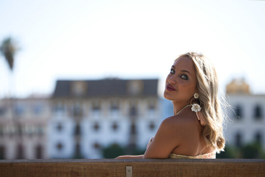 Young beautiful blonde woman sitting on a bench by the river bank in seville. The woman is relaxed and resting and looks back for the photo. Photo taken from behind.