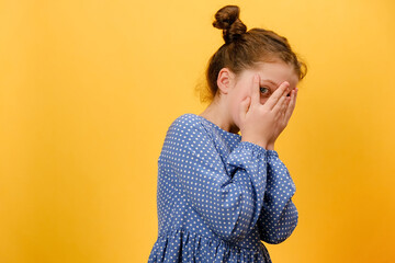 Portrait of shocked little girl kid covers face with hands, posing isolated over yellow background,...