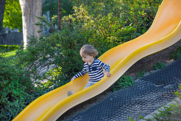 Happy cute little child boy having fun on a yellow slide outdoor in the park, sunny summer day in...