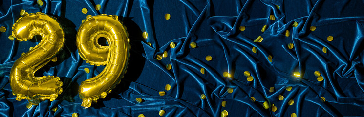 number twenty nine, gold foil balloon number on blue velvet background with confetti. Birthday greeting card, inscription 29. Anniversary event. Banner. Stylish gold numeral, bright shiny shine.