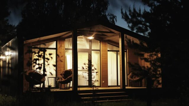 Night shot of wooden scandinavian house the light is on in the window, cozy terrace in summer with warm electric light. Forest cabin in the middle of a forest in silence, night sky, slow motion.