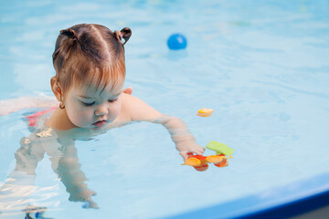 Fototapeta na wymiar Happy baby girl swims and plays with balls in pool with trainer. Concept developing fine motor skills and health care of child