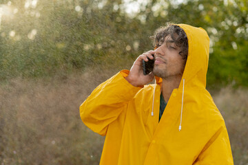 Young arabic man wearing a yellow raincoat calling on phone during the rain in the park. Guy...