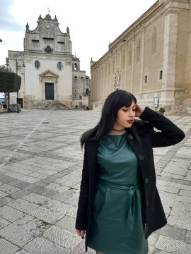 Brunette girl in a dark green dress, akin to petrol green, knee-high boots through the streets of Gravina di Puglia. Urban style and tradition merge in a vibrant image.