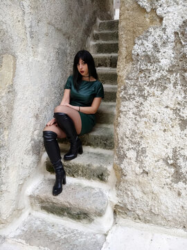 Brunette girl in a dark green dress, akin to petrol green, knee-high boots through the streets of Gravina di Puglia. Urban style and tradition merge in a vibrant image.
