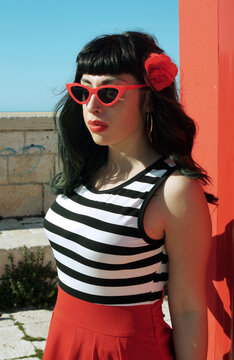 Port harbor pin-up: Red skirt, stripes, retro glasses. Red heels, intriguing secrets beneath: garter belt and sheer stockings. Revisited vintage elegance.