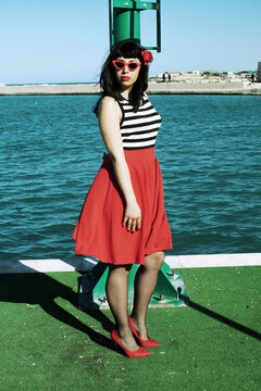 Port harbor pin-up: Red skirt, stripes, retro glasses. Red heels, intriguing secrets beneath: garter belt and sheer stockings. Revisited vintage elegance.