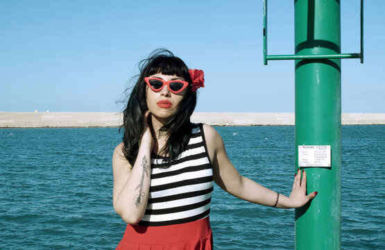 Port harbor pin-up: Red skirt, stripes, retro glasses. Red heels, intriguing secrets beneath: garter belt and sheer stockings. Revisited vintage elegance.