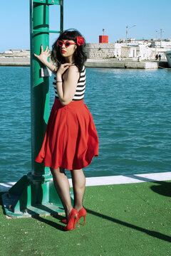 Port harbor pin-up: Red skirt, stripes, retro glasses. Red heels, intriguing secrets beneath: garter belt and sheer stockings. Revisited vintage elegance.
