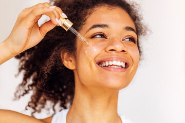 Cute smiling african young woman applies moisturizing serum from pipette to her cheek.