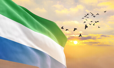 Waving flag of Sierra Leone against the background of a sunset or sunrise. Sierra Leone flag for Independence Day. The symbol of the state on wavy fabric. - obrazy, fototapety, plakaty