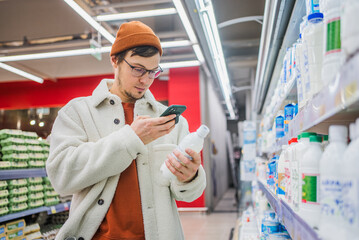 Man in store takes picture of label using phone. Young bearded Caucasian guy in glasses in department with dairy products and eggs holds kefir in hands, takes photo of composition, comparing.