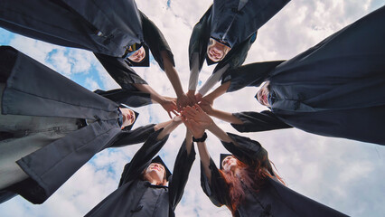 Group of happy successful graduates in academic hats and robes standing in circle and putting their hands together. Team of college or university students celebrating graduation.