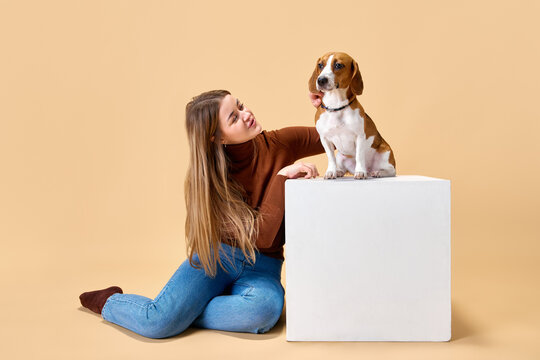 Portrait of young pretty woman together with her purebred Beagle, dog against beige studio background. Concept of animal, pet lover, friendship, domestic life, companionship. Ad. Copy space.