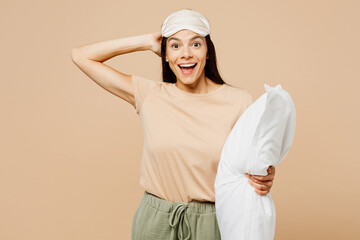 Young happy surprised calm Latin woman wears pyjamas jam sleep eye mask rest relax at home hold pillow look camera isolated on plain pastel light beige background studio. Good mood night nap concept.