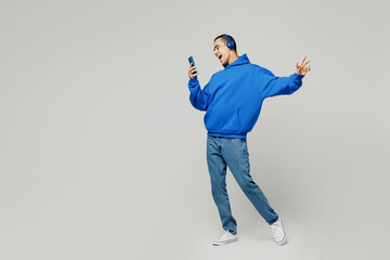Full body young middle eastern man he wears blue hoody casual clothes listen to music in headphones use mobile cell phone isolated on plain solid white background studio portrait. Lifestyle concept.