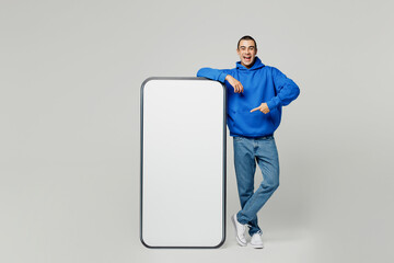 Full body young middle eastern man he wears blue hoody casual clothes point finger on big huge blank screen mobile cell phone with workspace copy space area isolated on plain solid white background.