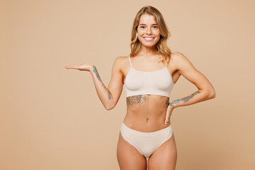 Young smiling nice lady woman with slim body perfect skin wear nude top bra lingerie stand akimbo point hand aside on area isolated on plain pastel light beige background. Lifestyle diet fit concept.