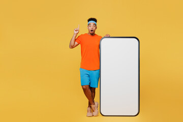 Full body young trainer sporty man sportsman wear orange t-shirt big huge screen mobile cell phone point finger up train in home gym isolated on plain yellow background. Workout sport fit abs concept.