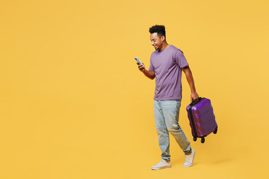 Traveler man wears casual clothes hold bag suitcase use mobile cell phone isolated on plain yellow background. Tourist travel abroad in free spare time rest getaway. Air flight trip journey concept.