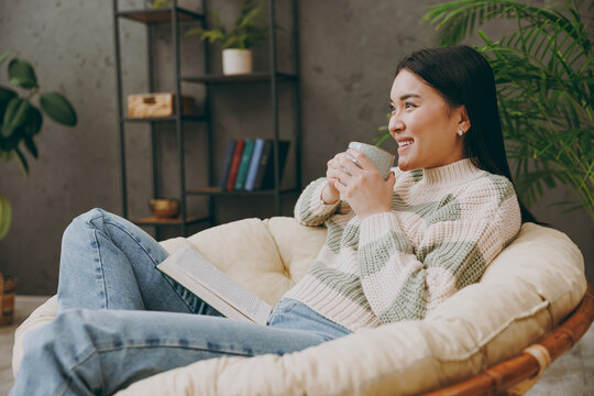 Side view young happy minded woman wear casual clothes sits on armchair read book novel drink tea stay at home hotel flat rest relax spend free spare time in grey living room indoor. Lounge concept.