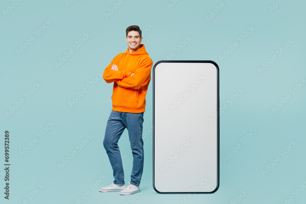 Wall mural Full body happy young man he wears orange hoody casual clothes stand near big huge blank screen mobile cell phone smartphone with area isolated on plain blue cyan color background. Lifestyle concept.