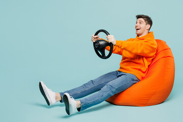 Full body young man he wears orange hoody casual clothes sit in bag chair hold steering wheel driving car isolated on plain pastel light blue cyan color background studio portrait. Lifestyle concept.